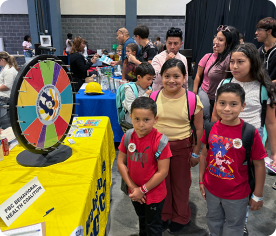 children smiling and standing in front of the PBCBHC table at a community event